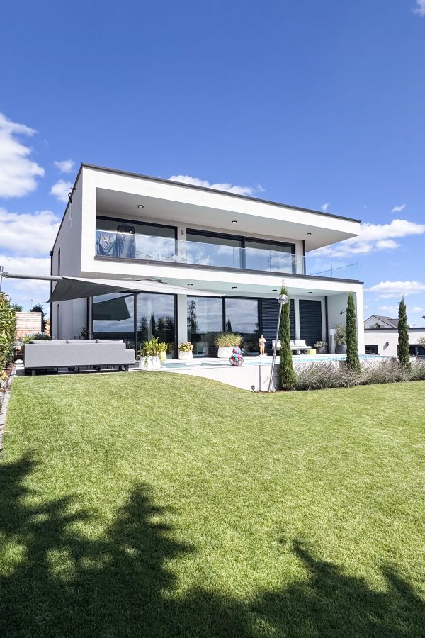 Moderne Rückansicht eines Einfamilienhauses mit großem Garten, Pool, Terrasse, Glasbalkon und Flachdacharchitektur.