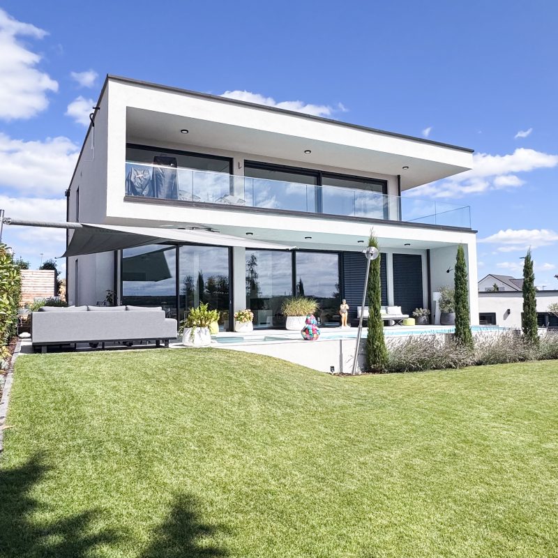 Moderne Rückansicht eines Einfamilienhauses mit großem Garten, Pool, Terrasse, Glasbalkon und Flachdacharchitektur.