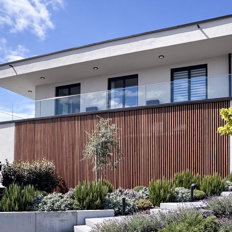 Moderne Hausfassade mit vertikaler Holzlamellenverkleidung, Glasbalkon und klarer Flachdacharchitektur.