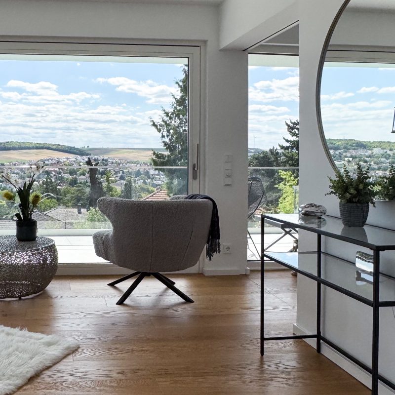 Heller Bad- und Loungebereich mit großem Panoramafenster, Sessel, rundem Spiegel und weitem Ausblick ins Umland.