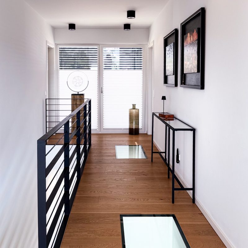 Moderner Flur im Obergeschoss mit Glasbodenelementen, schwarzem Geländer, Holzfußboden und klarer Linienführung.