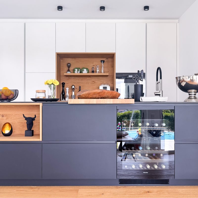 Moderne Küche mit Kücheninsel, dunklen Fronten, Holzakzenten, integriertem Weinkühlschrank und maßgefertigter Hochschrankwand.