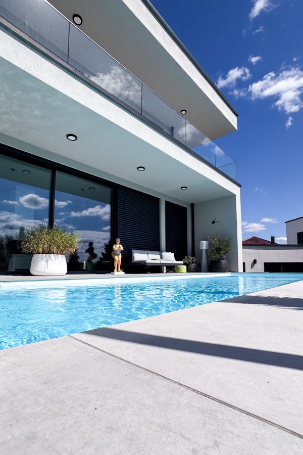 Moderne Rückansicht eines Einfamilienhauses mit Pool, überdachter Terrasse, Glasbalkon und großzügigen Fensterflächen.