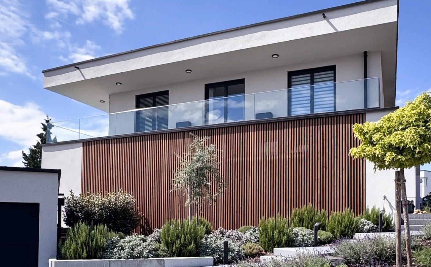 Moderne Hausfassade mit vertikaler Holzlamellenverkleidung, Glasbalkon und klarer Flachdacharchitektur.